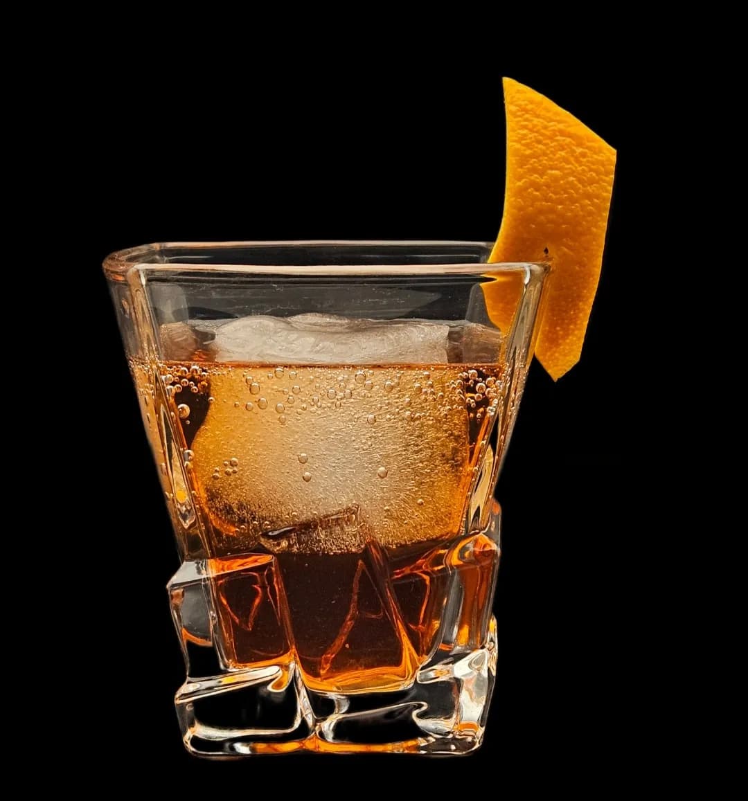 An amber cocktail over ice with an orange twist, lit against black